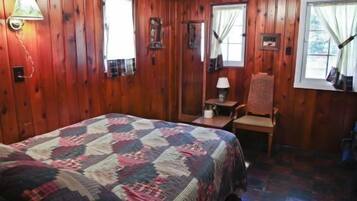 A wooden-paneled room with a bed, a chair, a table, and two windows.