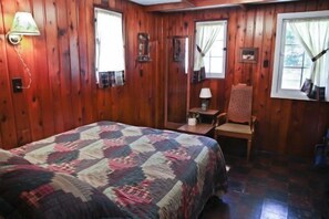 A wooden-paneled room with a bed, a chair, a table, and two windows. - #4 - Loon's Landing 2 Bedroom Cabin by RedAwning (Carp Lake)