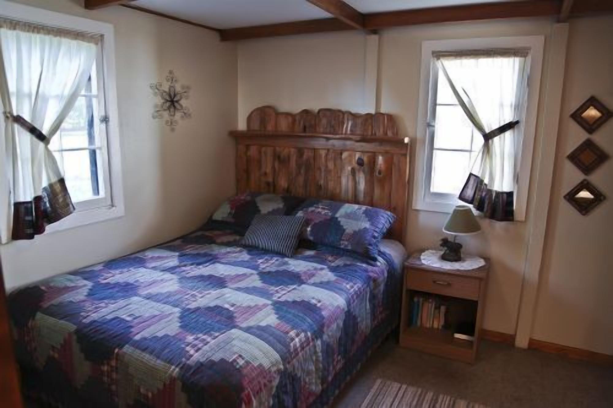 A bedroom with a wooden headboard, a quilted bedspread, a nightstand with a lamp, and a small bookshelf.