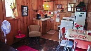 A compact kitchen with wooden walls, a dining area with a table and chairs, a small red chair, and a fan.