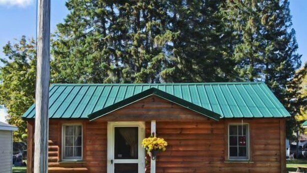 A small wooden cabin with a green roof, a front door, and a small porch area with a chair and a table.