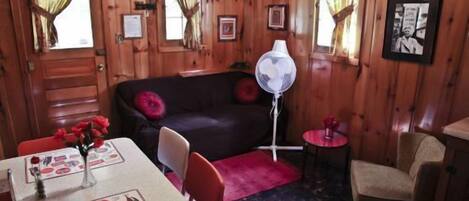 Interior de una cabaña de madera con un sofá negro, una mesa de comedor con un menú de Coca-Cola, un ventilador y una pequeña mesa redonda con un jarrón.