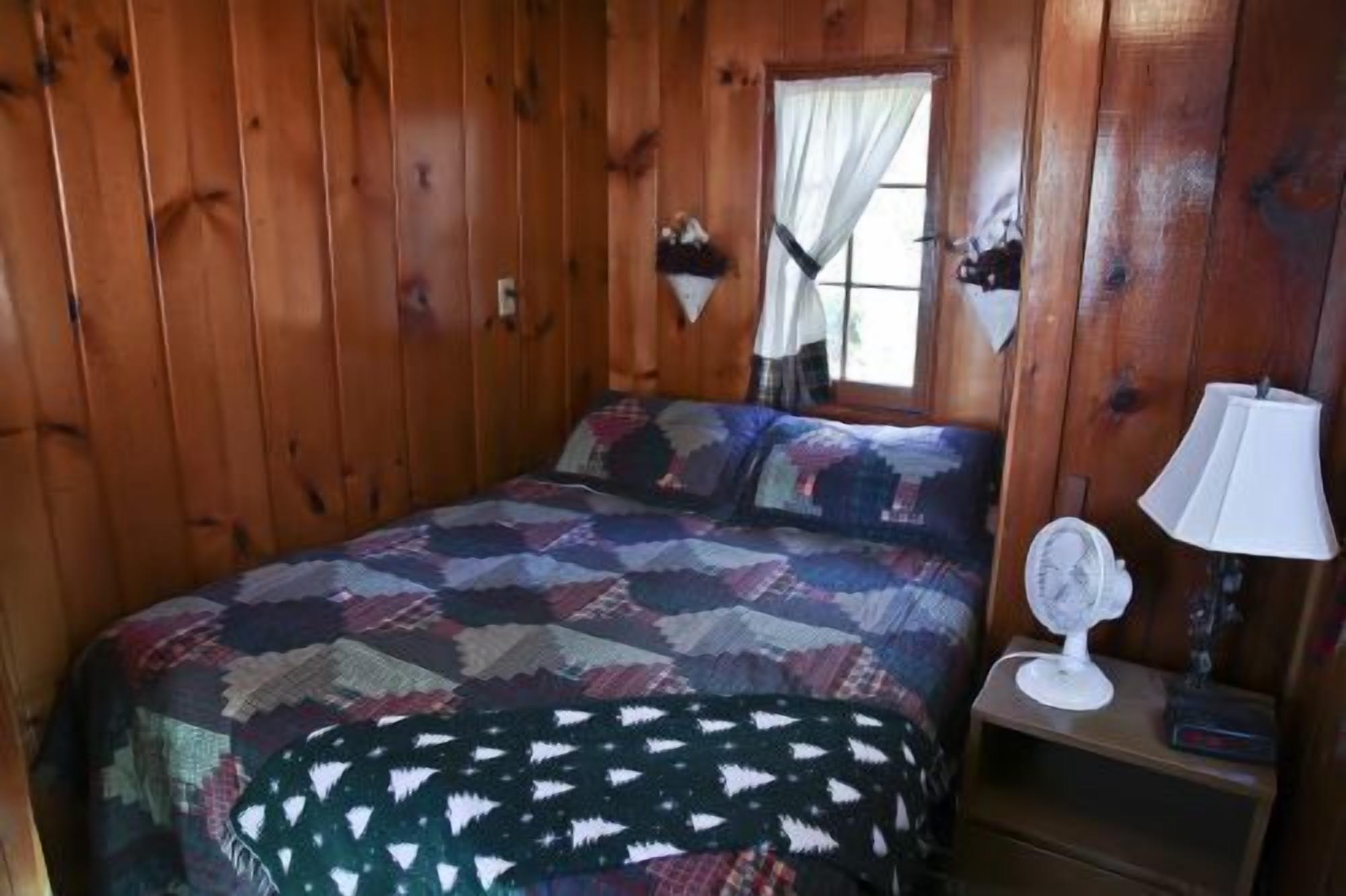 Un dormitorio de cabaña de madera con cama, mesita de noche, lámpara y ventana con cortinas.