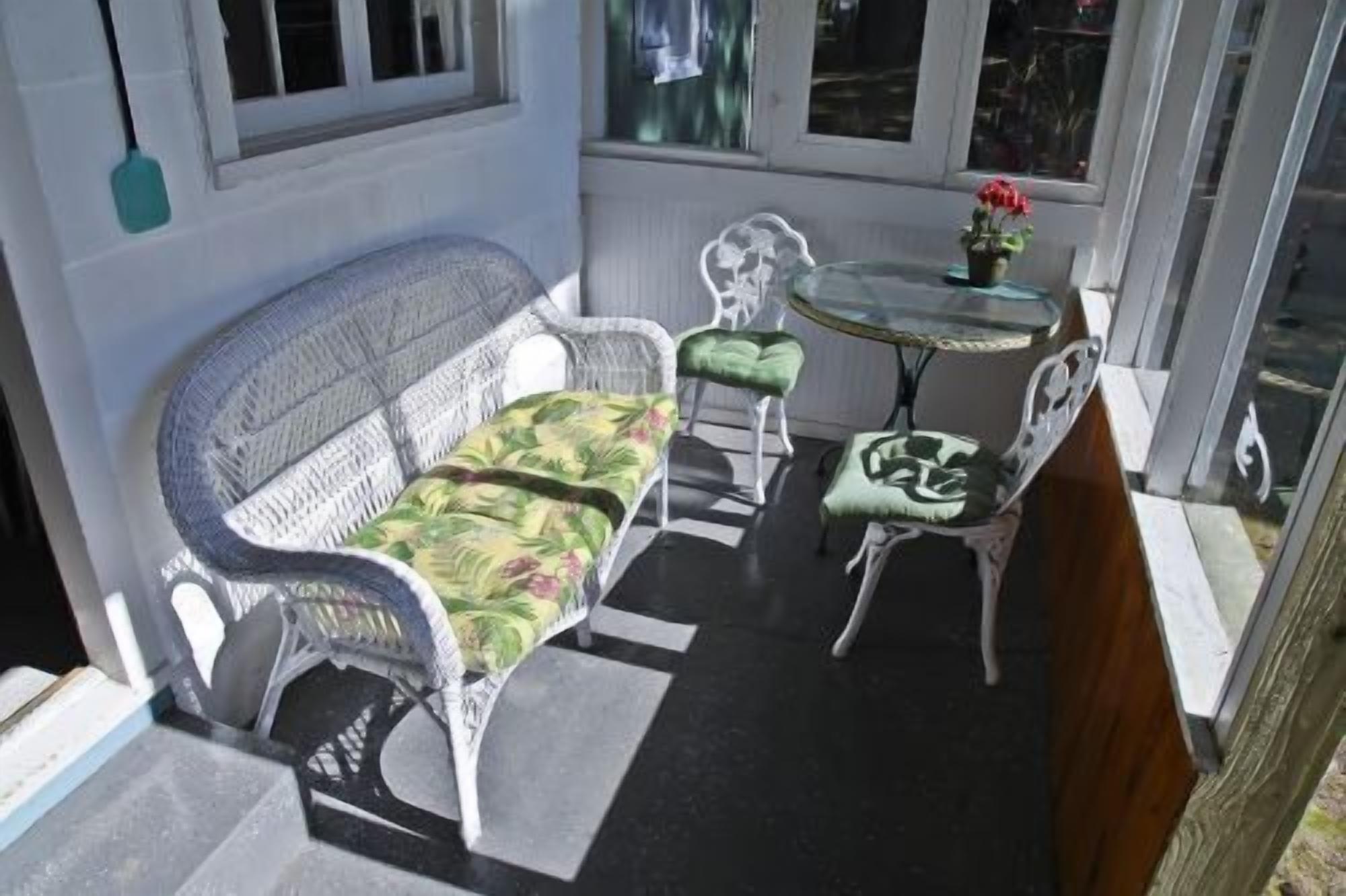 A sunlit patio with a white wicker sofa, two matching armchairs, a small round table, and a potted plant.