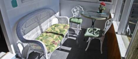 A sunlit patio with a white wicker sofa, two matching armchairs, a small round table, and a potted plant.