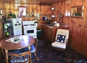 A compact kitchen with wooden walls, a dining table with chairs, and a small seating area with a cushioned chair. - #8 - Mackinac Point 1 Bedroom Cabin by RedAwning (Carp Lake)