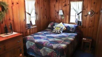 A wooden cabin bedroom with a bed, dresser, and two windows.