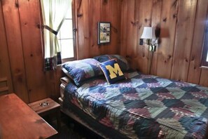 Habitación de cabaña de madera con una cama, una mesita, una lámpara y una ventana con cortinas.