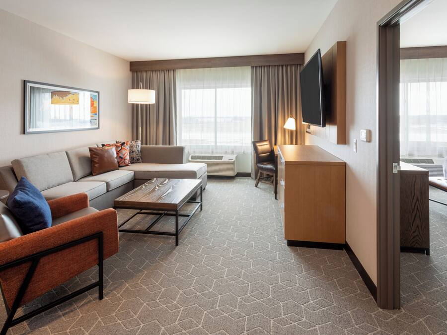 A hotel room with a sofa, armchair, and a small table. There is a TV mounted on the wall and a window with curtains.