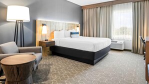 Habitación de hotel con cama, sofá, mesa redonda, lámpara, cabecero y ventana con cortinas.