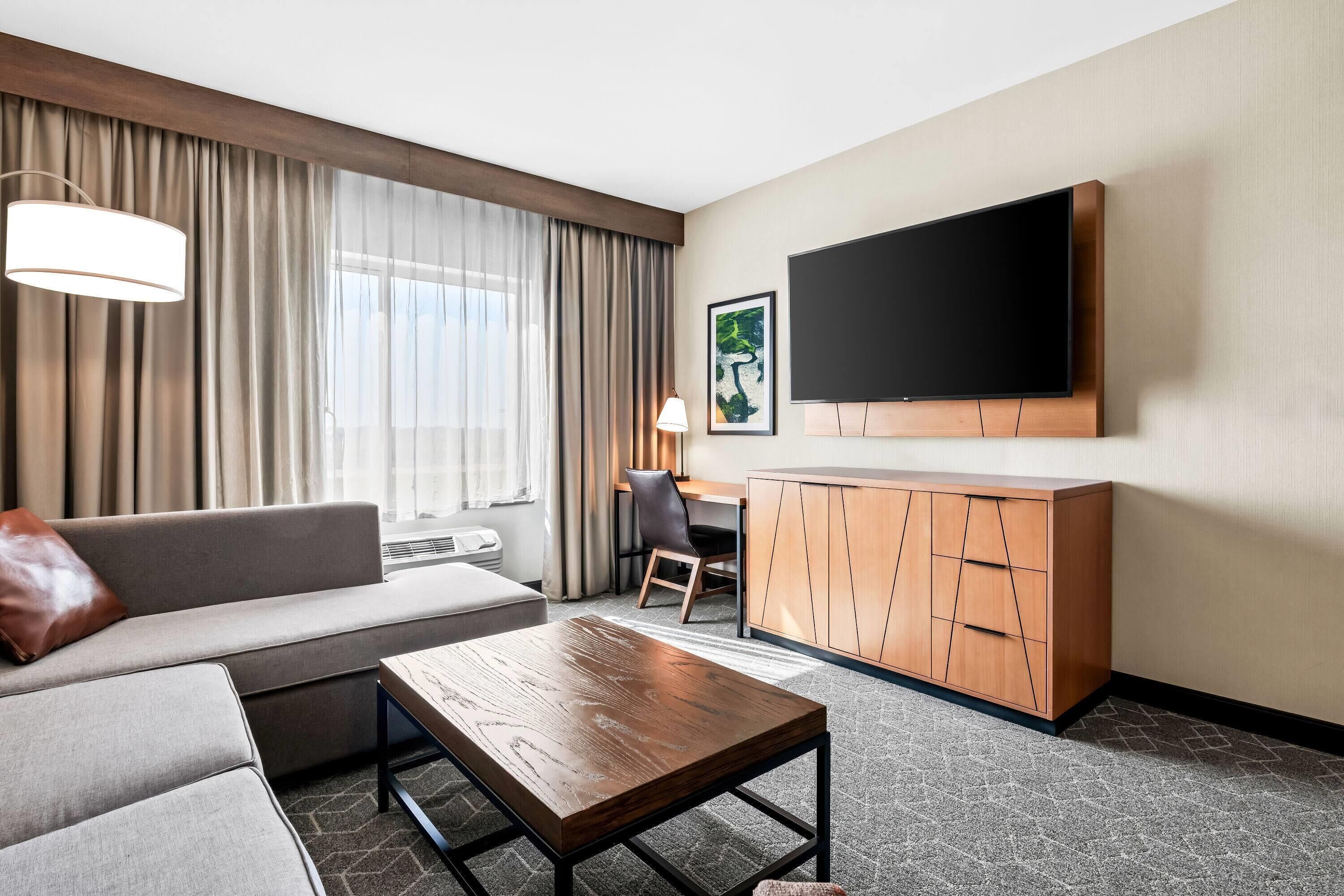 A modern hotel room with a sofa, a wooden coffee table, a desk, and a flat-screen TV mounted on the wall.