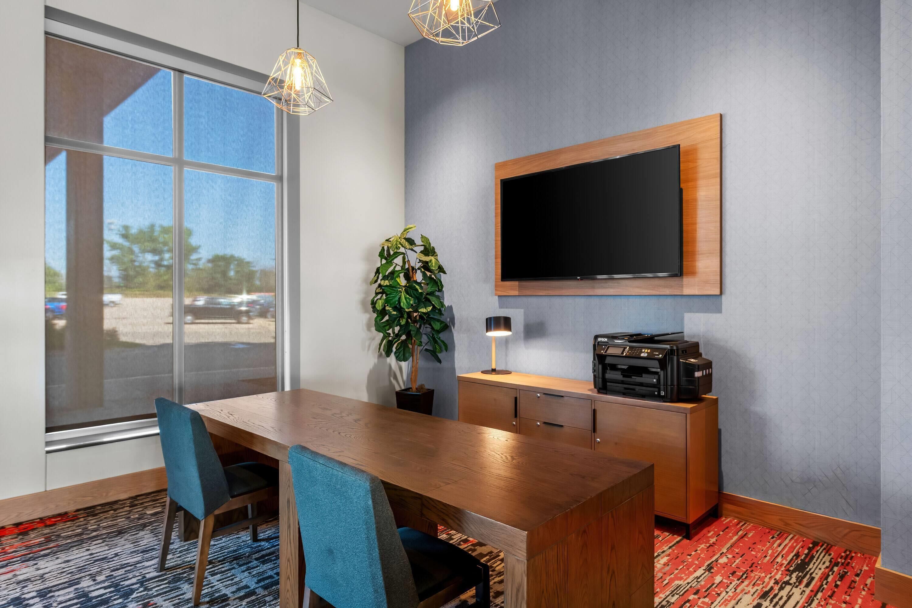 Habitación de hotel con un ventanal grande, una mesa de comedor de madera, dos sillas, un televisor montado en la pared, una planta e impresora sobre un mueble de madera.