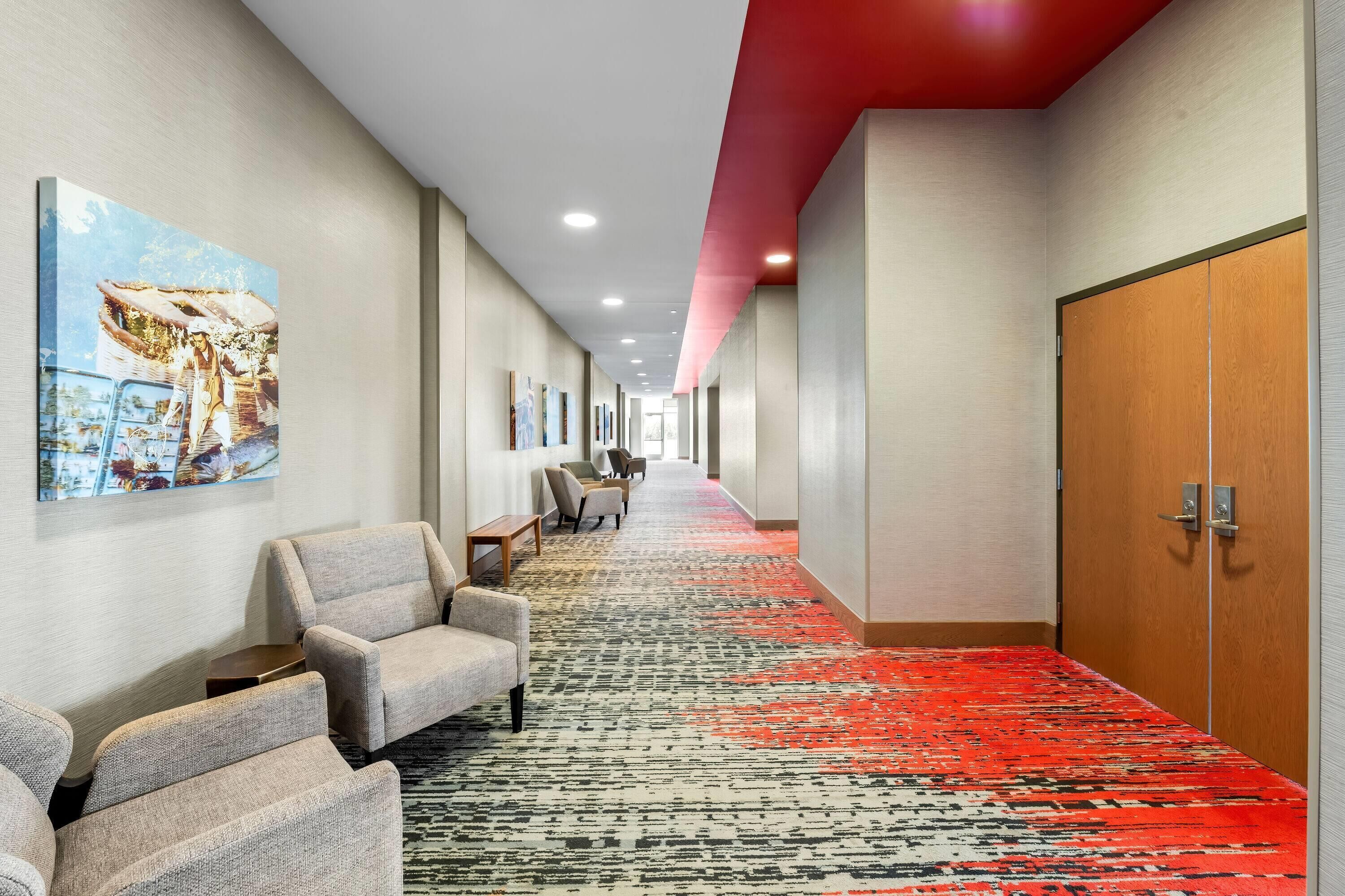 Un pasillo de hotel con alfombra roja, zona de estar con sillones y una puerta de madera.