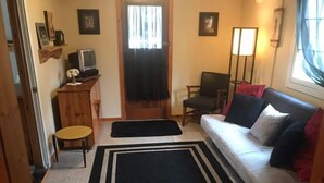 A living room with a sofa, a chair, a small table, a TV, and a door leading to another room. - #5 - Times Square 2 Bedroom Cabin by RedAwning (Carp Lake)