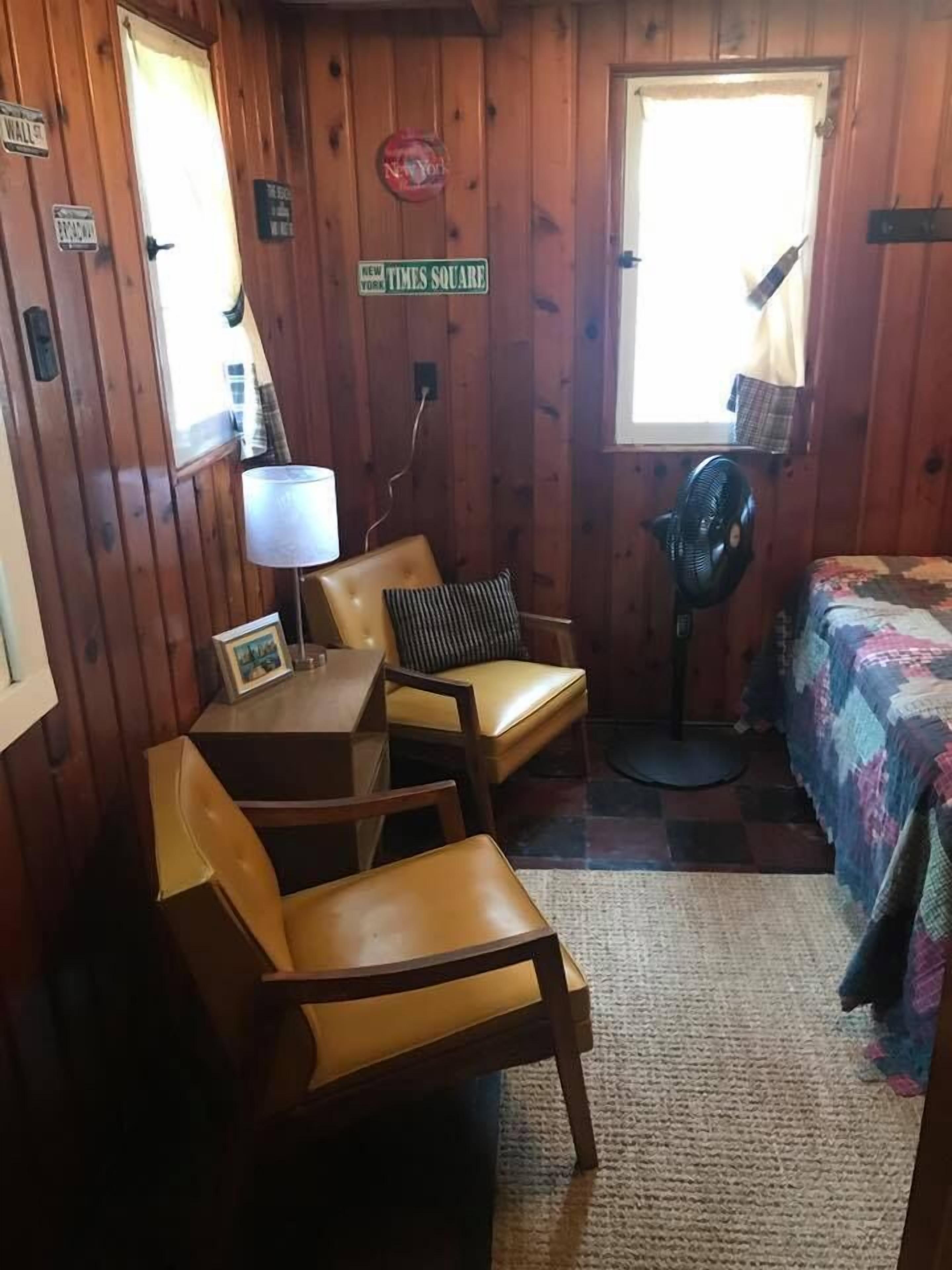 A small, cozy room with wooden walls, a bed with a colorful quilt, two chairs, a small table with a lamp, and a fan.