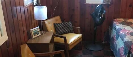 A small, cozy room with wooden walls, a bed with a colorful quilt, two chairs, a small table with a lamp, and a fan.