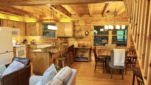 A cozy cabin-style kitchen with wooden cabinets, a central island, and a dining area with a table and chairs. - Lake Hideaway 2 - Lakeside Log Cabin, Fishing Boat, Private Dock & Mountain View (Cherry Log)