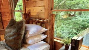 A cozy cabin interior with a bed, pillows, a window with a view of a forest, and a small table with books and a plant. - Lake Hideaway 2 - Lakeside Log Cabin, Fishing Boat, Private Dock & Mountain View (Cherry Log)