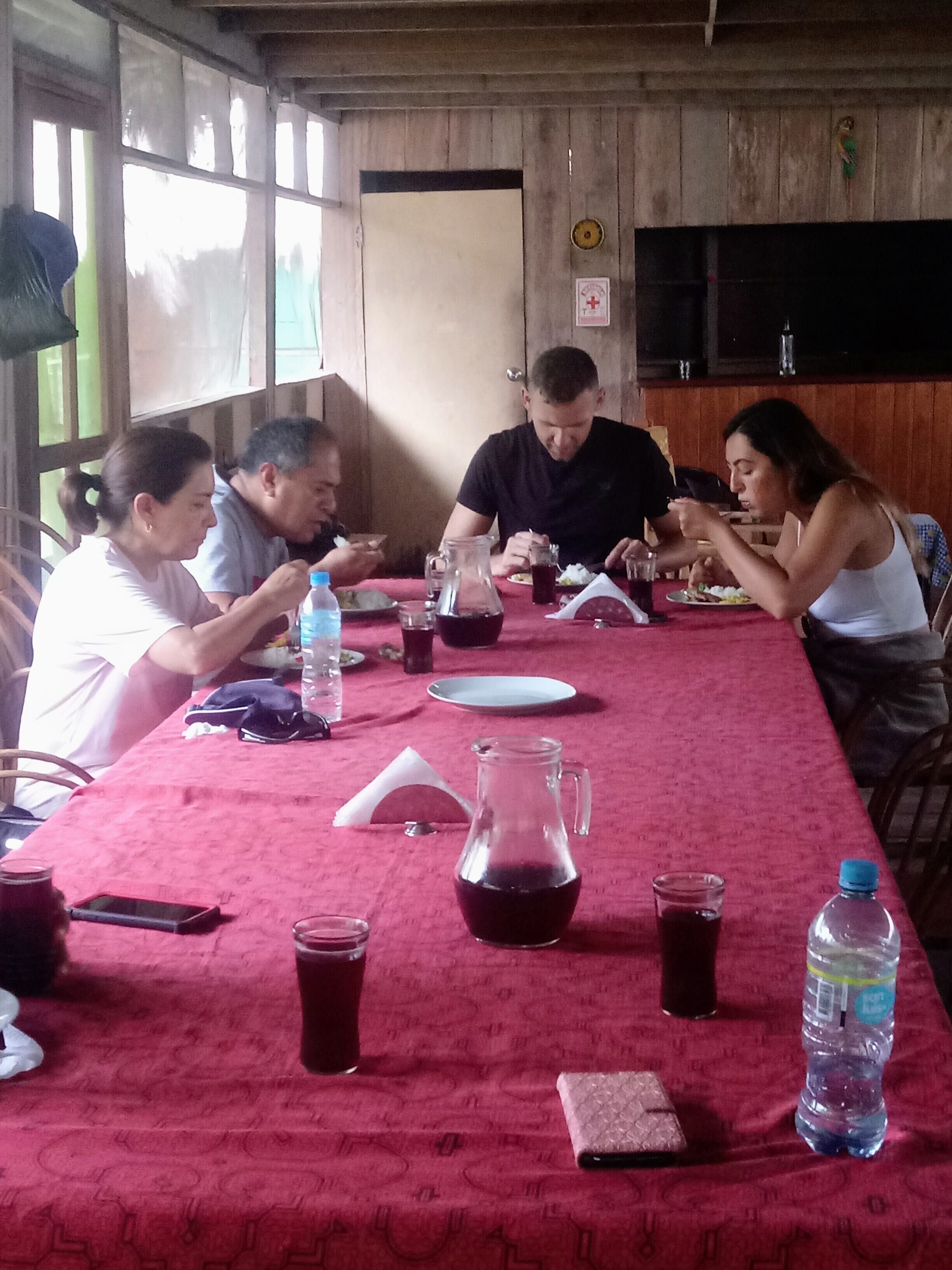 Un gruppo di persone cena a un tavolo con una tovaglia rossa, diversi piatti e bevande.