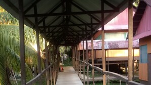 A covered walkway with a wooden floor, surrounded by greenery and buildings with corrugated metal roofs. - Ecoadventure Amazon (Indiana)