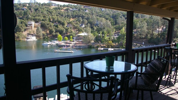 Un espace extérieur aménagé, abrité et doté de tables et de chaises, offrant une vue sur un lac où l’on peut apercevoir des bateaux et des maisons sur une colline.