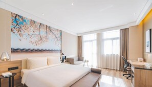 Une chambre d’hôtel moderne avec un grand lit, un bureau et une chaise. Il y a un tableau au mur et une fenêtre avec des rideaux.