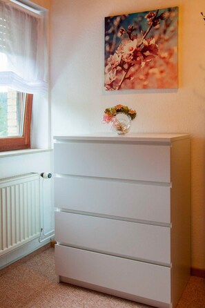 Ein weißer Kommode mit Schubladen, ein Fenster mit durchsichtigen Vorhängen und ein an der Wand hängendes Bild von Blumen.