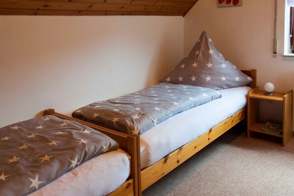 Ein kleines Zimmer mit einem Einzelbett, einem Holzbettgestell, einem Nachttisch, einer Lampe und einem Fenster.