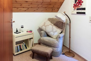 Ein gemütliches Zimmer mit Holzbalkendecke, einem gepolsterten Sessel, einem Schreibtisch mit Büchern, einer Lampe und einem Wandbild.