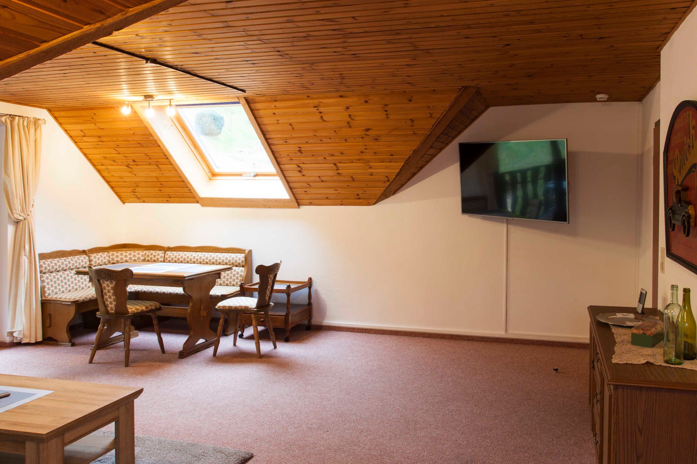 Une pièce avec un plafond en pente, une table à manger en bois avec des chaises, un téléviseur fixé au mur et un petit meuble contenant des bouteilles.