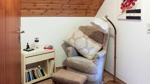 Una habitación acogedora con techo de madera, un sillón mullido, un escritorio con libros, una lámpara y una obra de arte en la pared.
