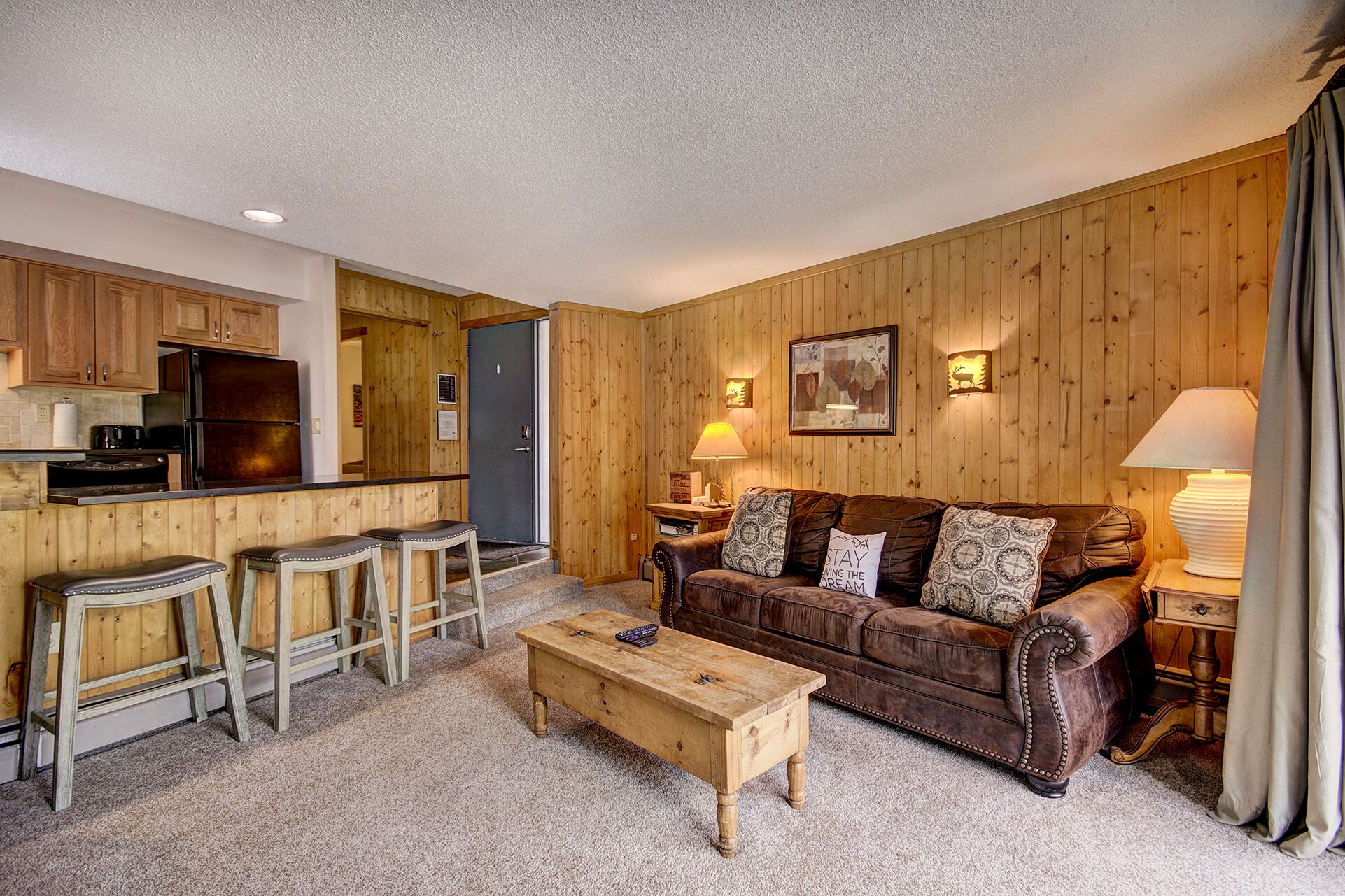 A cozy living room with a brown leather sofa, wooden coffee table, and a kitchen area with wooden cabinets and a refrigerator.
