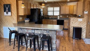 Eine Küche mit Holzschränken, einer Granit Arbeitsplatte und einer Wand mit Natursteinverkleidung. Drei schwarze Barhocker und ein schwarzer Kühlschrank sind vorhanden.