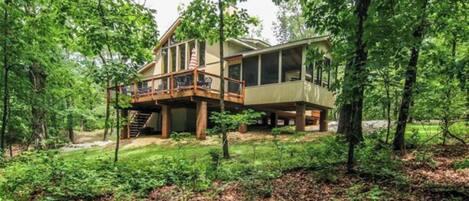 A two-story wooden cabin with a deck, surrounded by dense forest.