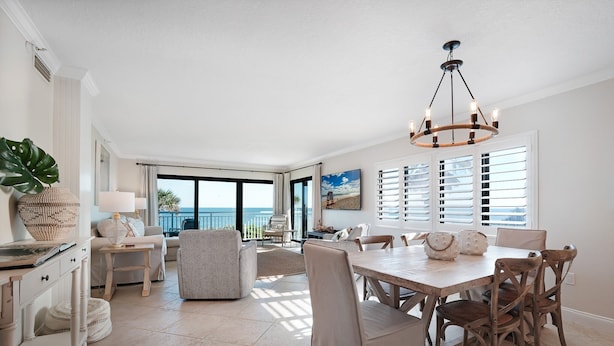 A spacious living room with a large window overlooking the ocean, a dining area with wooden furniture, and a kitchen with white cabinetry. - Direct oceanfront 2/2 corner condo with large wrap-around balcony and heated pool (New Smyrna Beach)