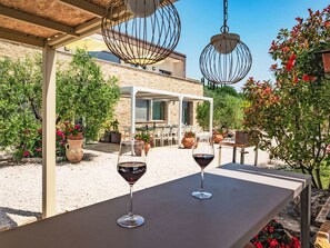Un área de estar al aire libre con dos copas de vino sobre una mesa, rodeada de macetas con plantas y un edificio de piedra al fondo.