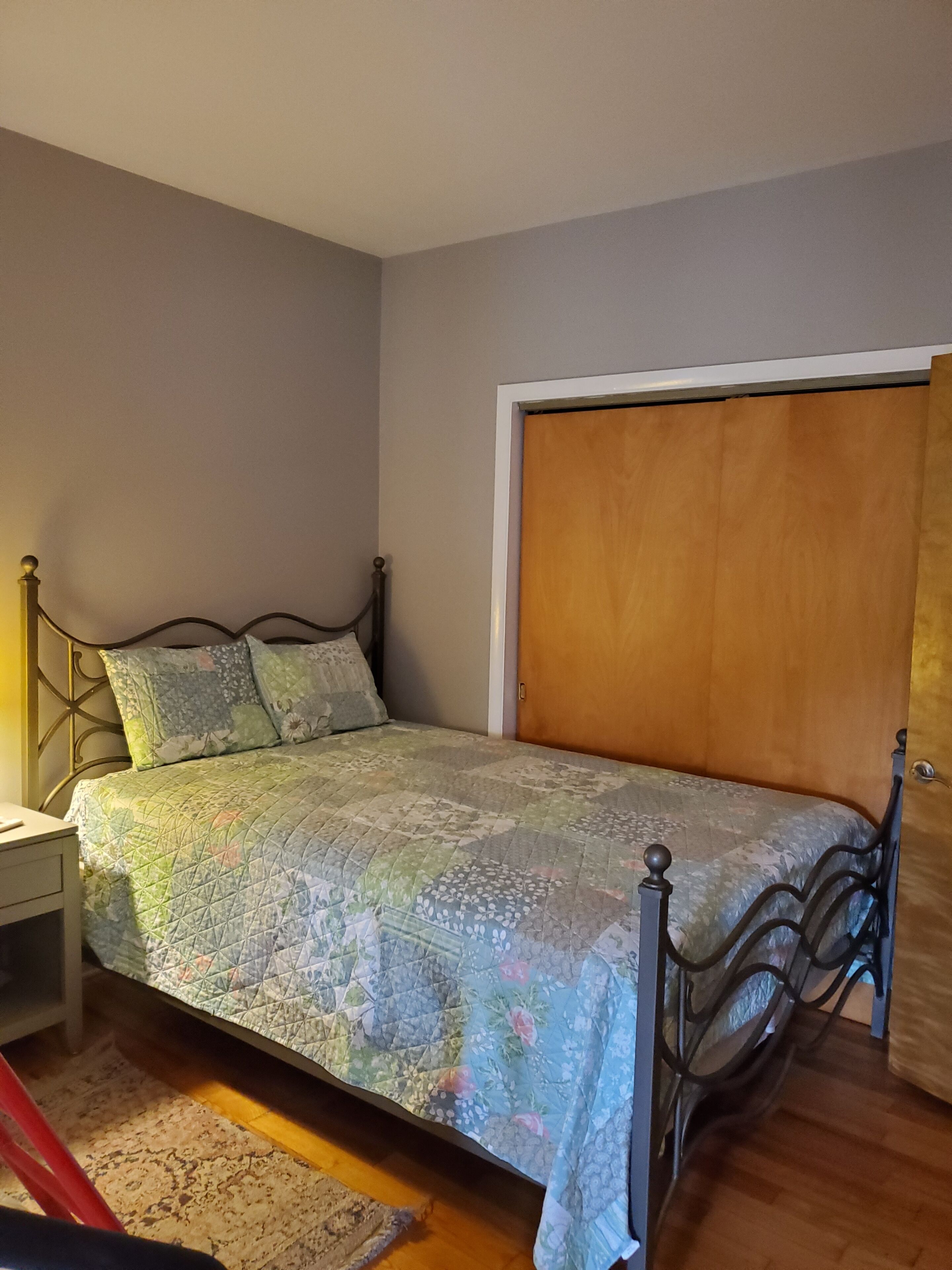 Quarto com cama, mesa de cabeceira, porta de madeira e uma cadeira vermelha.