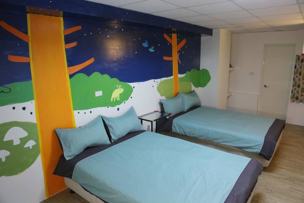 A room with two beds, each with a blue bedspread and matching pillows. The walls are painted with a nature-themed mural, including trees and a moon.