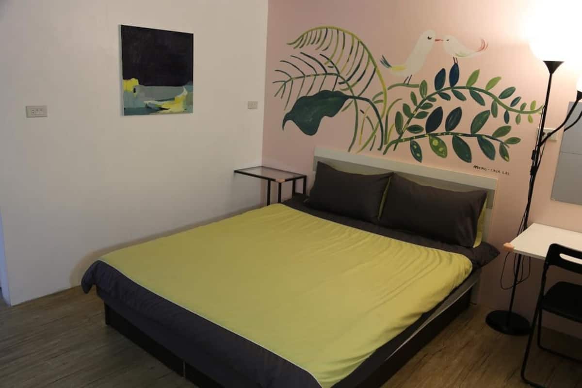 A bedroom with a bed, a nightstand, a desk, and a chair. There is a wall with a painting of birds and leaves.