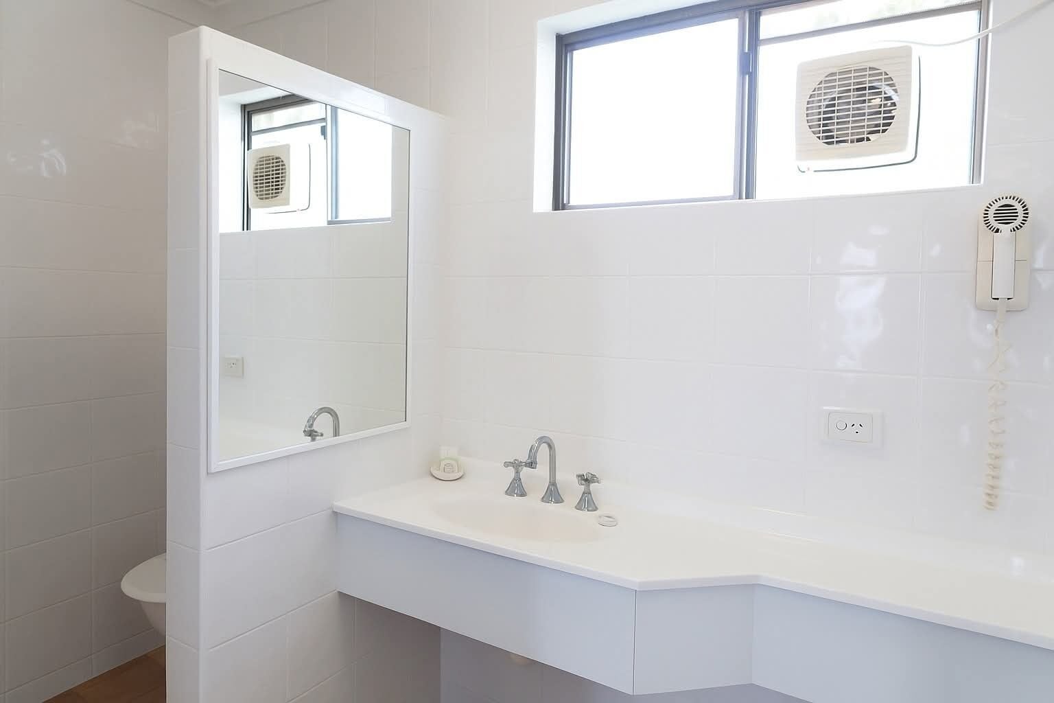 A modern bathroom with a white sink, two faucets, a mirror, a window with a fan, and a hairdryer.