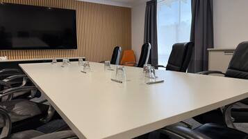 Un salón de conferencias con una larga mesa, numerosas sillas, un televisor de pantalla plana grande y un ventanal con cortinas.