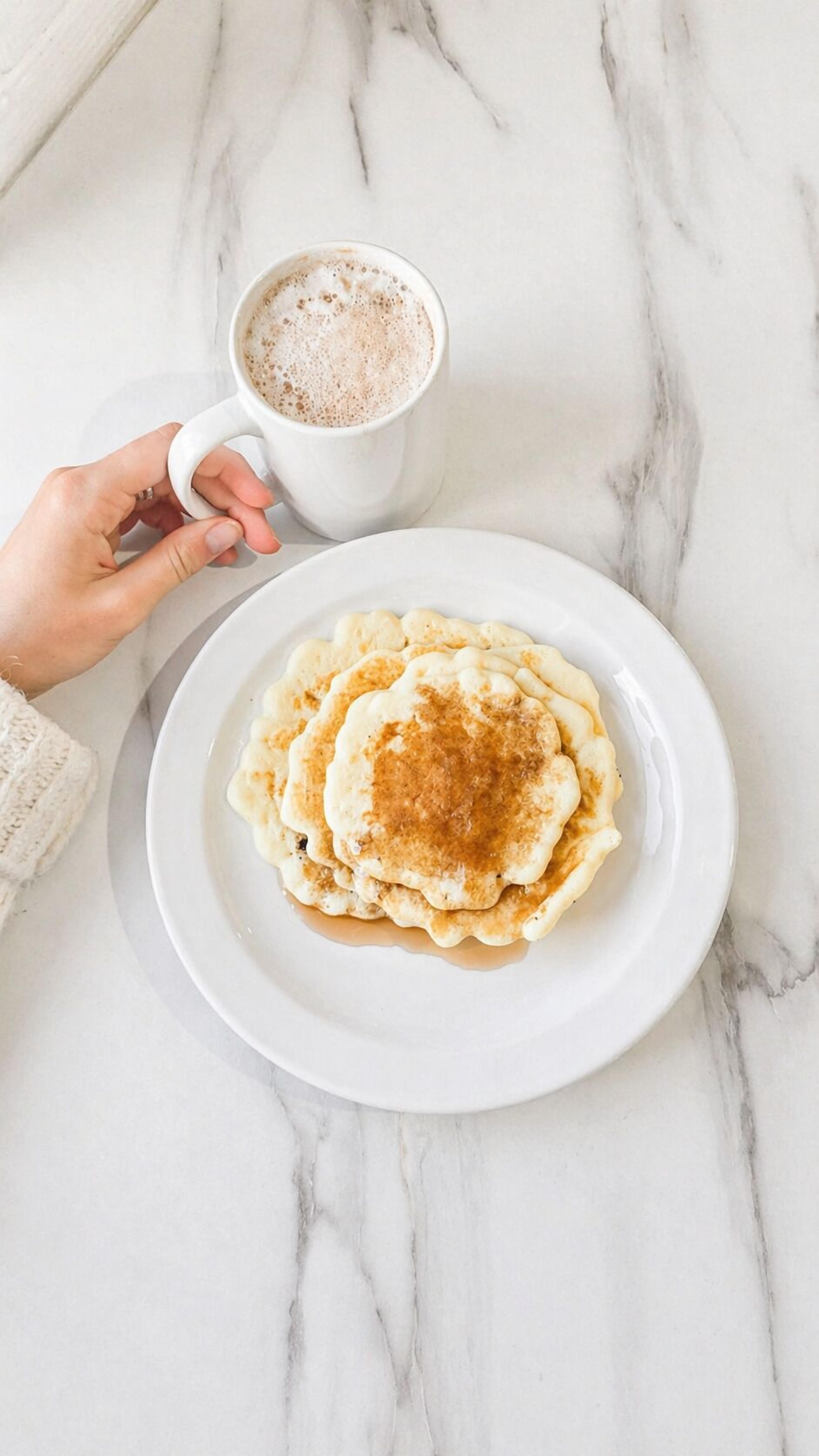 A white marble surface with a white plate of pancakes and a white cup of coffee.