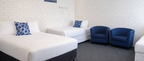Habitación de hotel con dos camas, un sillón azul, un sillón blanco y un cuadro en la pared.