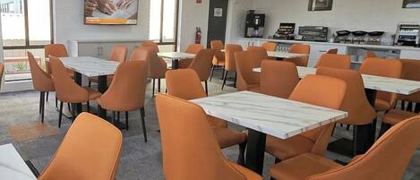 A modern dining area with orange chairs and white tables, equipped with an air conditioning unit and a coffee machine.