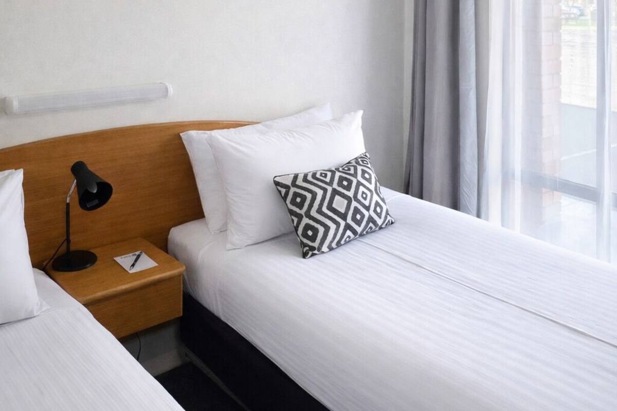 A hotel room with a single bed, a wooden headboard, a bedside lamp, and a window with curtains.