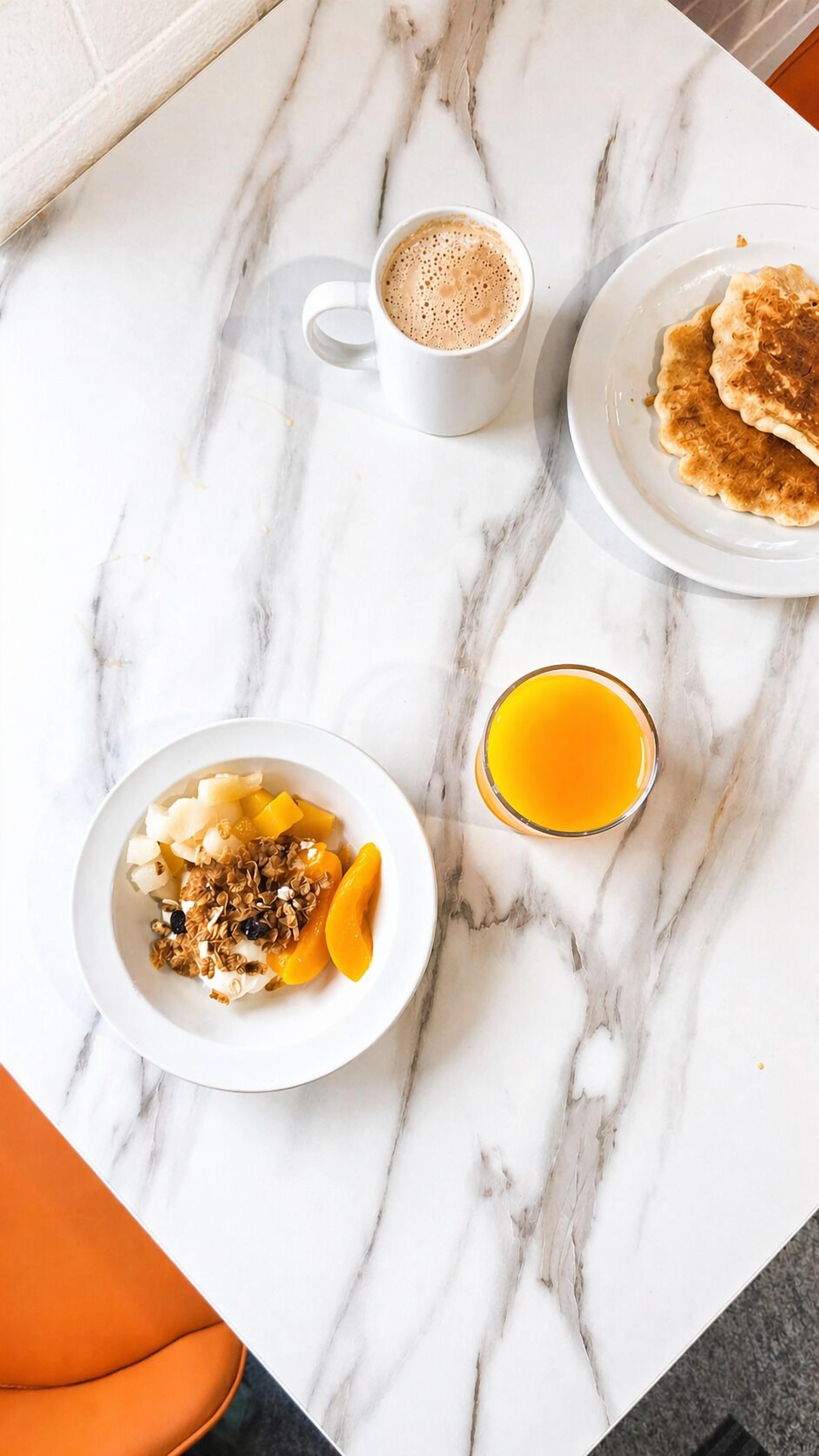 Ein Frühstückstisch mit Marmorplatte, eine Tasse Kaffee, Pfannkuchen, eine Schüssel Obst und Granola sowie ein Glas Orangensaft.