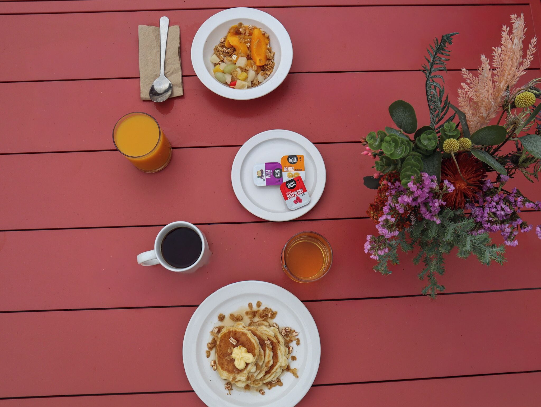 A breakfast setting with a plate of pancakes, a bowl of cereal with fruits, a glass of orange juice, a cup of coffee, a glass of tea, and a small plate with three packets of ZOOSH.