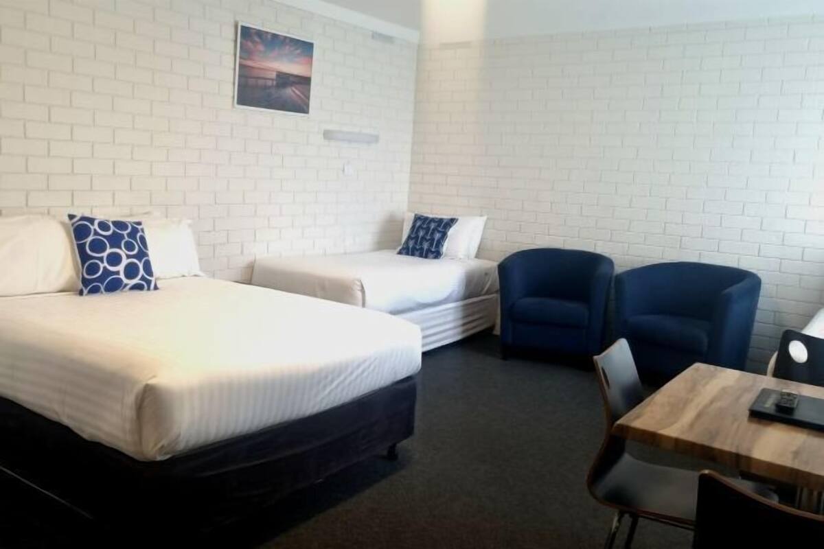 A room with two beds, a blue armchair, a wooden table, and a white brick wall.