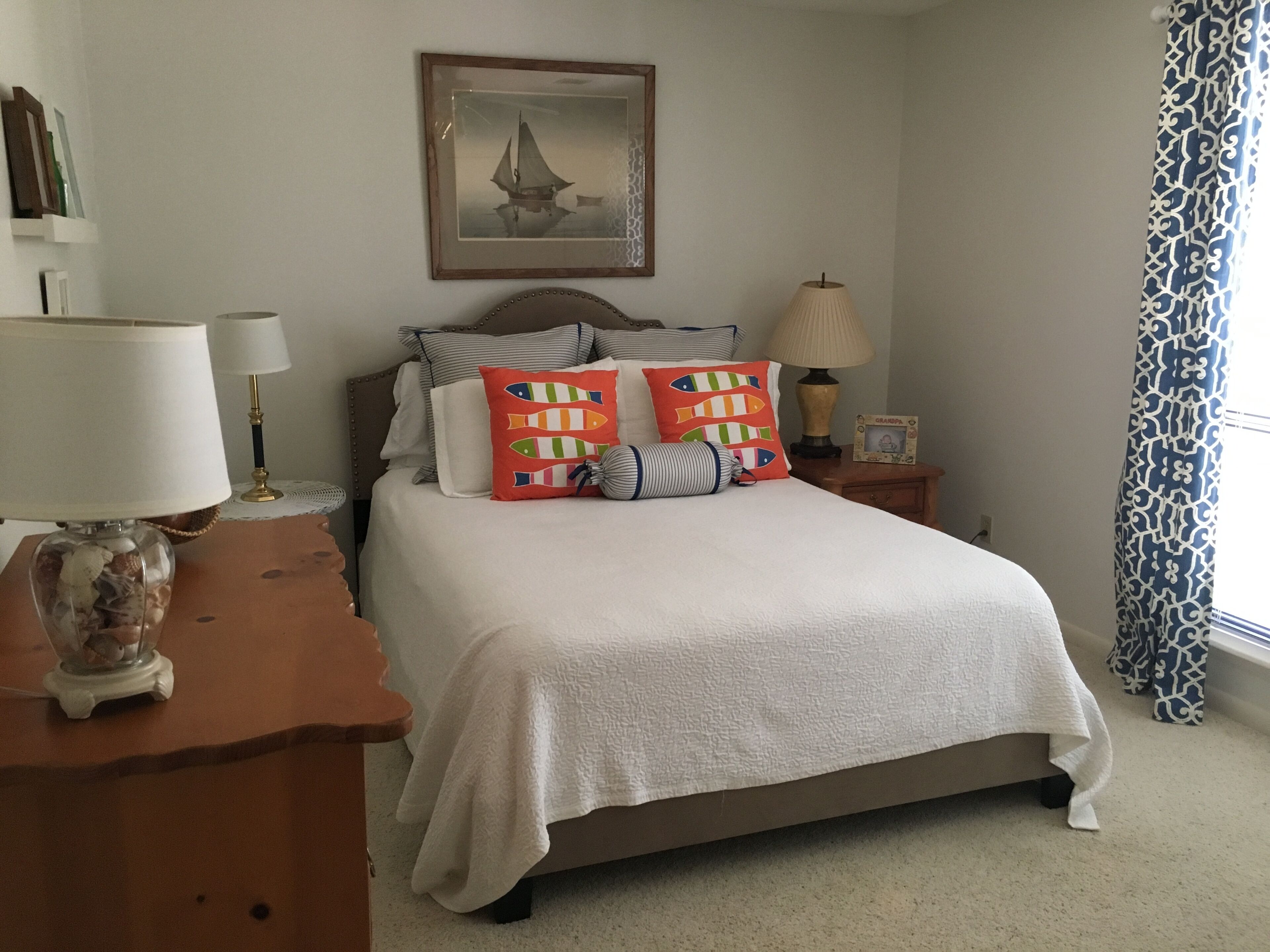 A neatly arranged bedroom with a bed, bedside tables, lamps, a framed picture on the wall, and a patterned curtain.