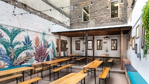An outdoor seating area with wooden tables and benches, a mural on the wall, and a brick building in the background.